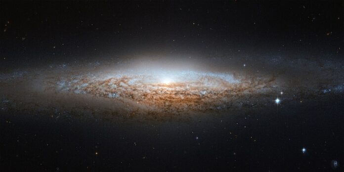 Edge on view of spiral galaxy NGC 2683, with a bright core and thin golden disk surrounded by faint blue star clouds against a dark space background.
